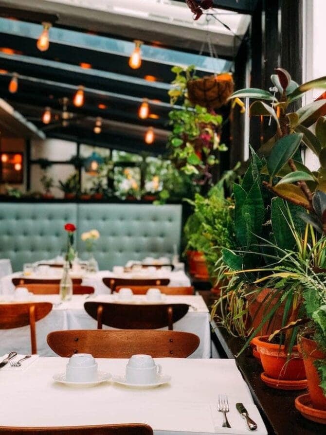 inside of a restaurant with white table clothes, wood chairs, booths, plants and edison lights.