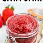 Glass jar full of strawberry chia jam. Jar is on a wood board, strawberries are in the background.