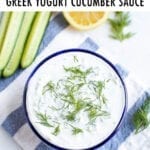 Bowl of tzatziki topped with fresh dill. The bowl is on a striped cloth and surrounded by cucumber, lemon, dill and a spoon.