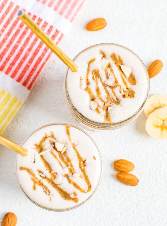 Bird's eye view of two banana almond butter smoothies with straws and topped with chopped almonds and an almond butter drizzle. Almonds, banana slices and a napkin are on the table.