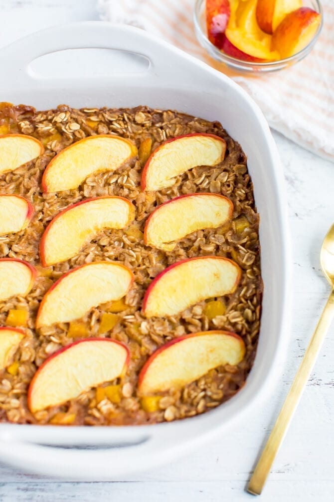 Baked oatmeal in a square dish and topped with peaches. A bowl of peach slices and a spoon are to the side.