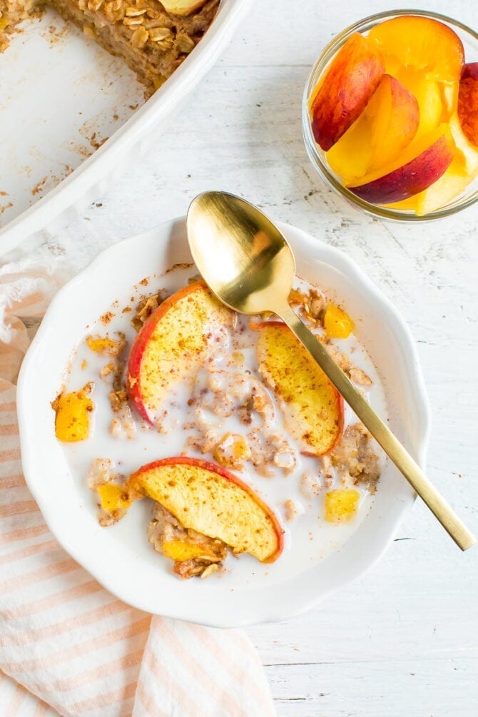 Bowl of peach baked oatmeal topped with cinnamon and almond milk. A spoon is resting on the bowl. A bowl of peaches and the baking dish are next to the bowl of oatmeal.