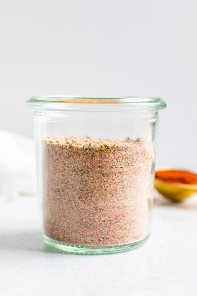 Glass jar with blackened seasoning. A teaspoon of paprika is in the background.
