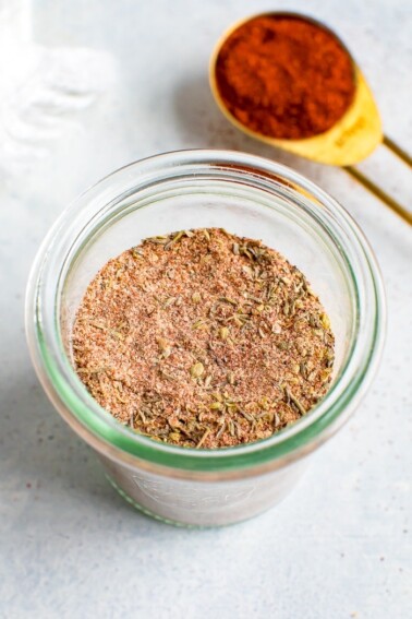 Glass jar with blackened seasoning. A teaspoon of paprika is in the background.