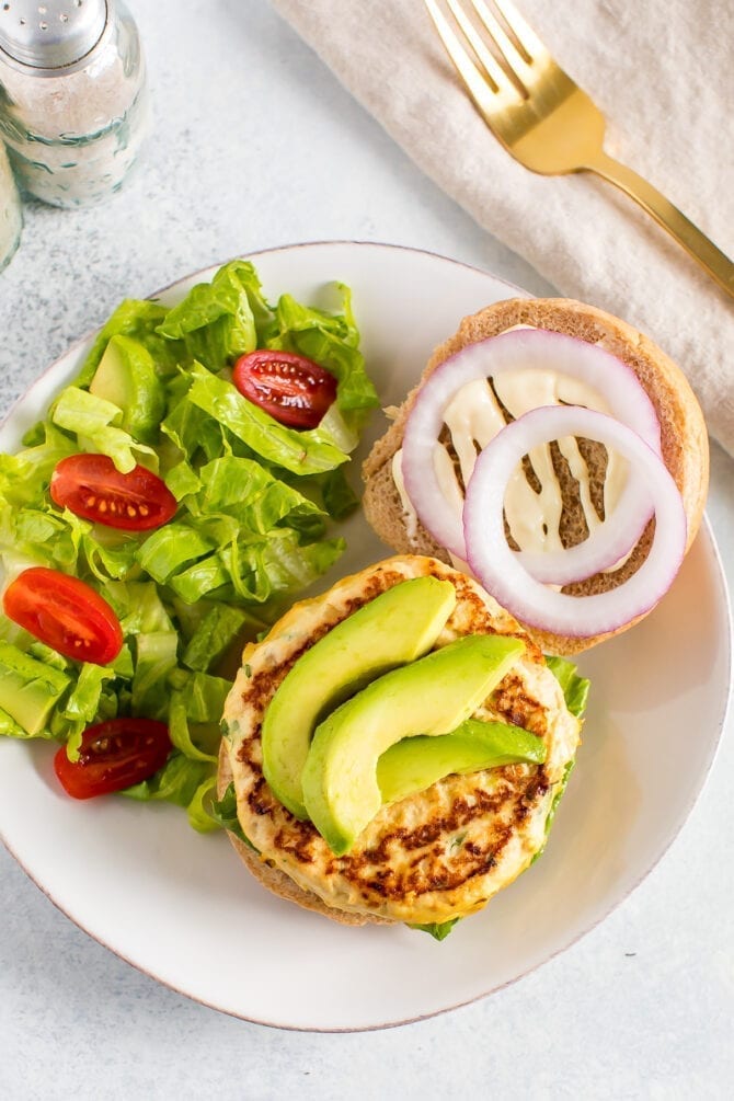 Chicken burger on an open face bun. Burger is topped with sliced avocado. Bun is topped with aioli and onions. A green salad is on the side of the plate.