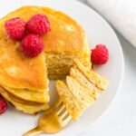 Stack of coconut flour pancakes topped with raspberries. A fork has taken a bite out of the stack.