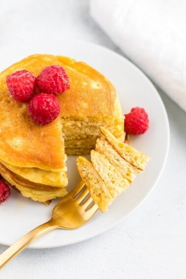 Stack of coconut flour pancakes topped with raspberries. A fork has taken a bite out of the stack.