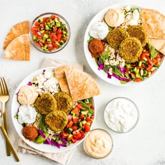 two falafel bowls on a table surrounded by cucumber tomato salad in a bowl, dips and pita.