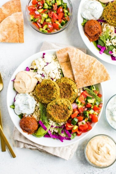 Falafel bowl on a table surrounded by cucumber tomato salad in a bowl, dips and pita.
