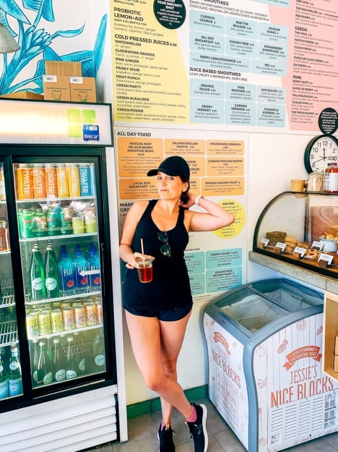 Woman wearing a baseball cap and holding an iced coffee, standing in a market.