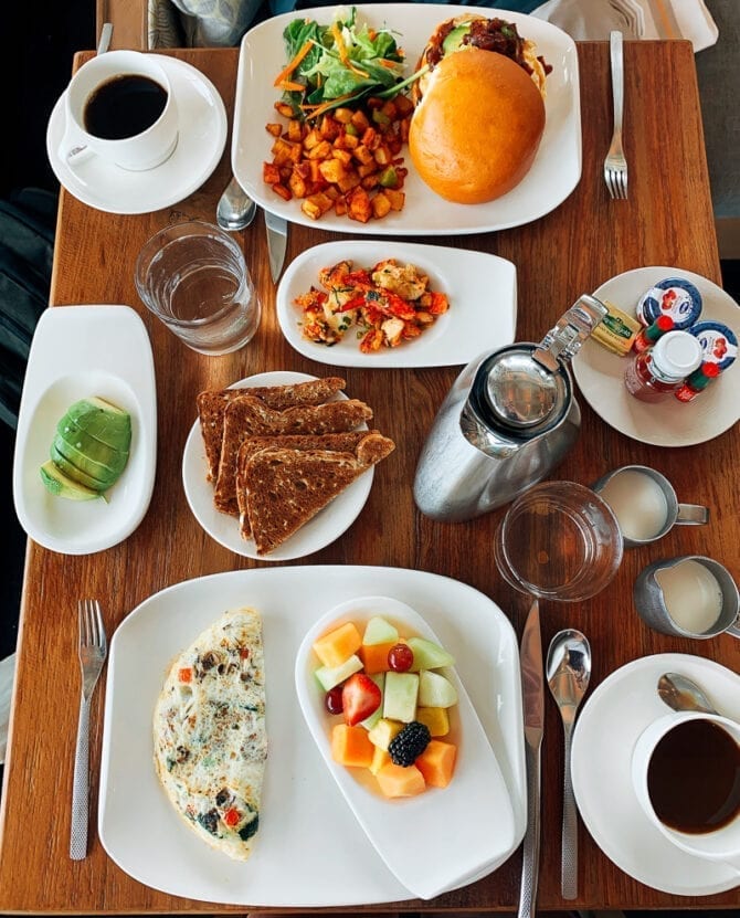 Restaurant table full of breakfast food-- breakfast sandwiches, hash, toast, eggs, and fruit.