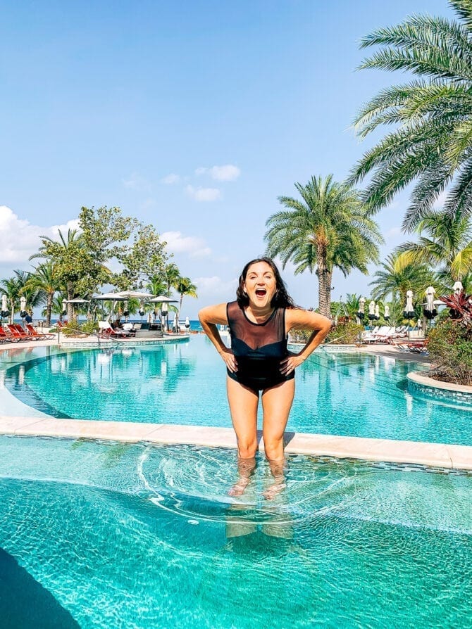 Woman in a black swimsuit standing in a Grand Cayman resort pool surrounded by palm trees.