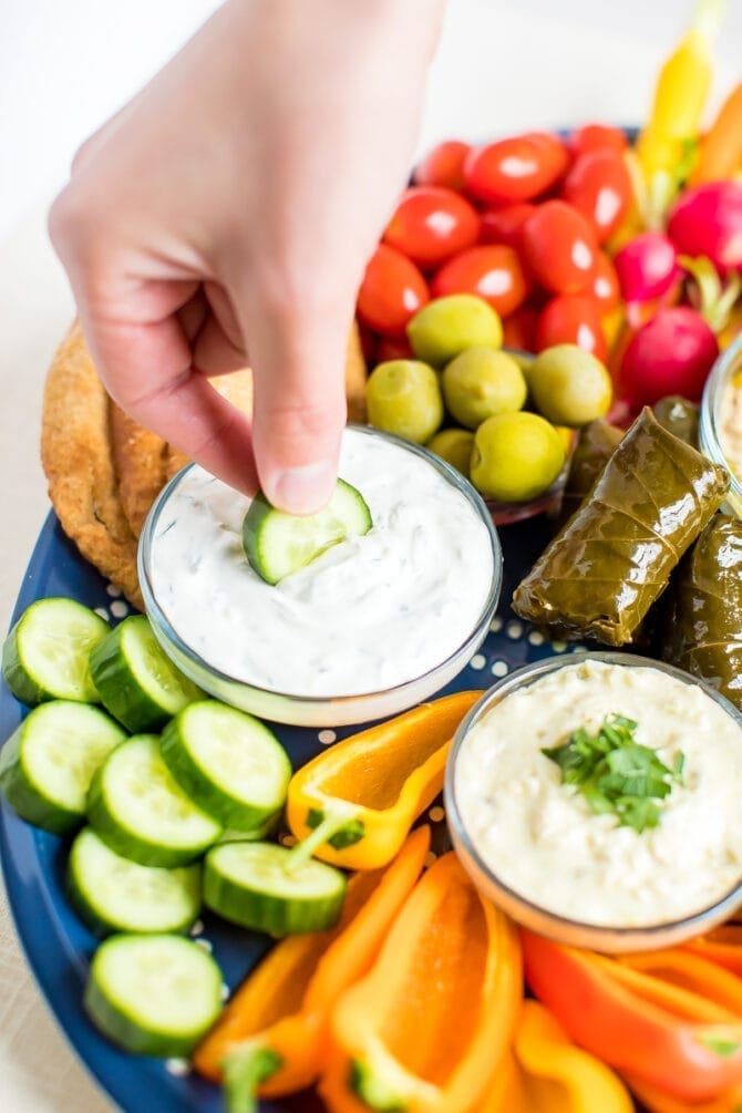 Hand dipping a cucumber into a bowl of tzaziki on a mezze platter.