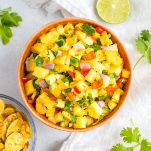 Bowl of pineapple salsa next to a bowl of plantain chips, cilantro sprigs and a slice of lime.
