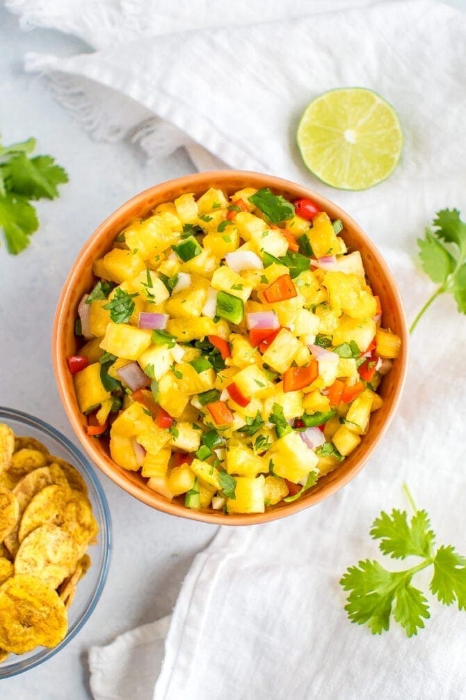 Bowl of pineapple salsa next to a bowl of plantain chips, cilantro sprigs and a slice of lime.