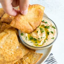 Hand dipping a pita chip into a bowl of hummus.