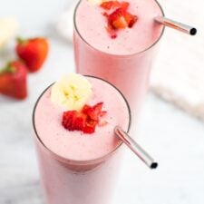 Two strawberry banana smoothies with a silver straw and topped with a banana slice and chopped strawberries.