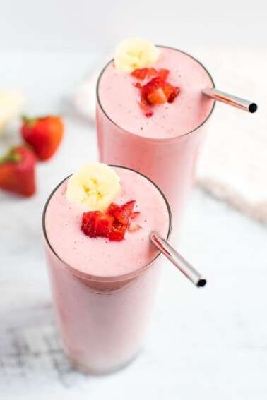 Two strawberry banana smoothies with a silver straw and topped with a banana slice and chopped strawberries.