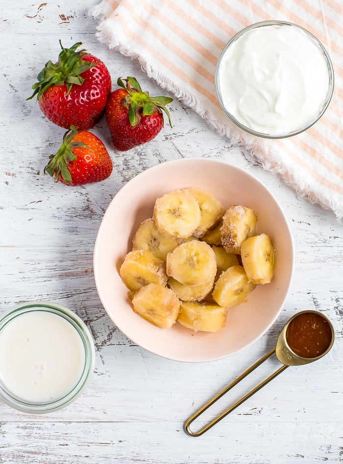 Ingredients to make a healthy strawberry banana smoothie. Fresh strawberries, a bowl of sliced frozen banana, a bowl of greek yogurt, a teaspoon of honey, and a jar of almond milk.