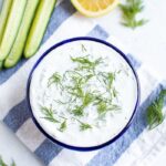 Bowl of tzatziki topped with fresh dill. The bowl is on a striped cloth and surrounded by cucumber, lemon, and dill.