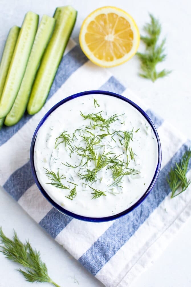 Bowl of tzatziki topped with fresh dill. The bowl is on a striped cloth and surrounded by cucumber, lemon, dill and a spoon.