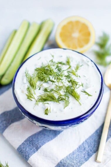 Bowl of tzatziki topped with fresh dill. The bowl is on a striped cloth and surrounded by cucumber, lemon, dill and a spoon.