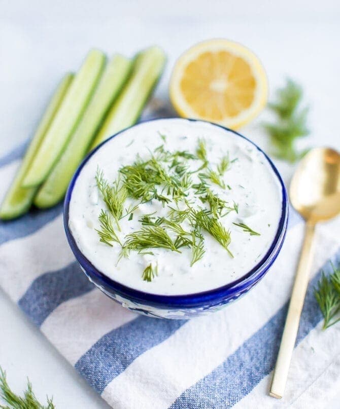 Bowl of tzatziki topped with fresh dill. The bowl is on a striped cloth and surrounded by cucumber, lemon, dill and a spoon.