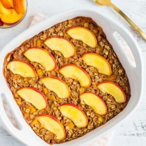 Peach baked oatmeal in a square dish and topped with peaches. A bowl of peach slices and a spoon are to the side.