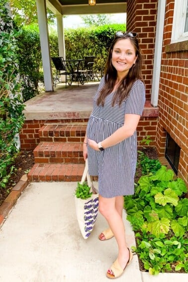 Pregnant woman in a striped dress smiling outside with a tote bag of vegetables.