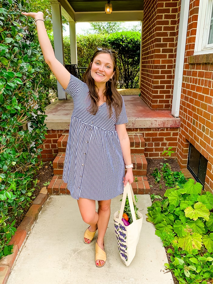 Woman in a striped dress smiling outside with a tote bag of vegetables.