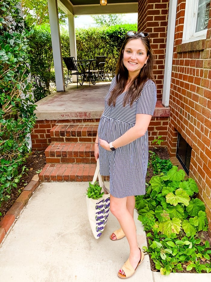 Pregnant woman in a striped dress smiling outside with a tote bag of vegetables.