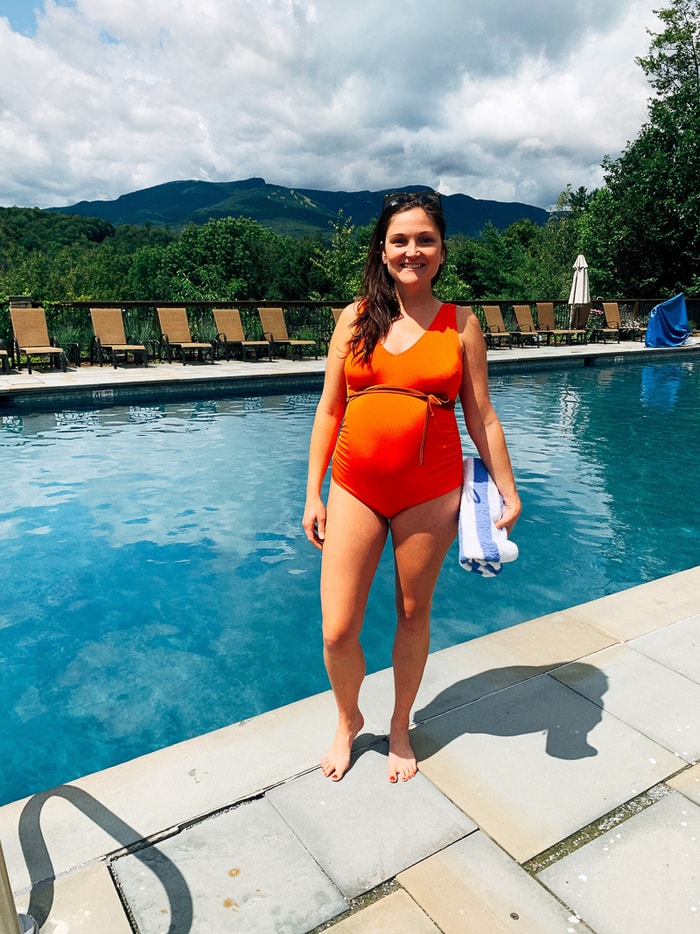 Woman in an orange swim suit standing in front of a pool with a view of the mountains.