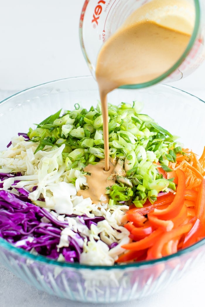 Asian dressing being poured into a bowl of the ingredients for an asian slaw before being tossed. Ingredients in the bowl are cabbage, scallions, carrots, and peppers.