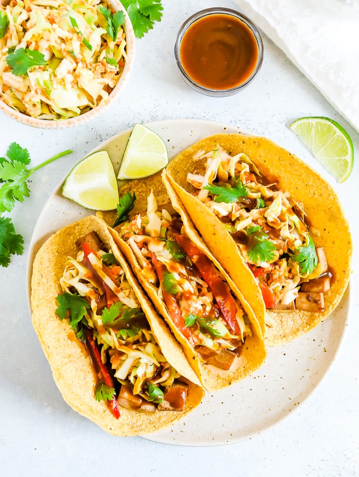 Three asian tofu tacos on a plate and served with lime. A bowl of slaw and teriyaki sauce are to the side of the place. Tacos are filled with slaw, teriyaki tofu, and cilantro.
