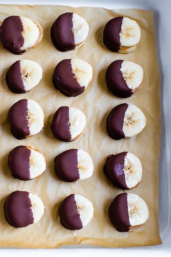 Slices of banana sandwiched with peanut butter, frozen, and half dipped with chocolate lined up on a tray lined with parchment paper.