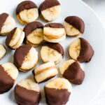 Chocolate peanut butter banana bites on a plate. Slices of banana sandwiched with peanut butter and dipped half in chocolate and frozen.