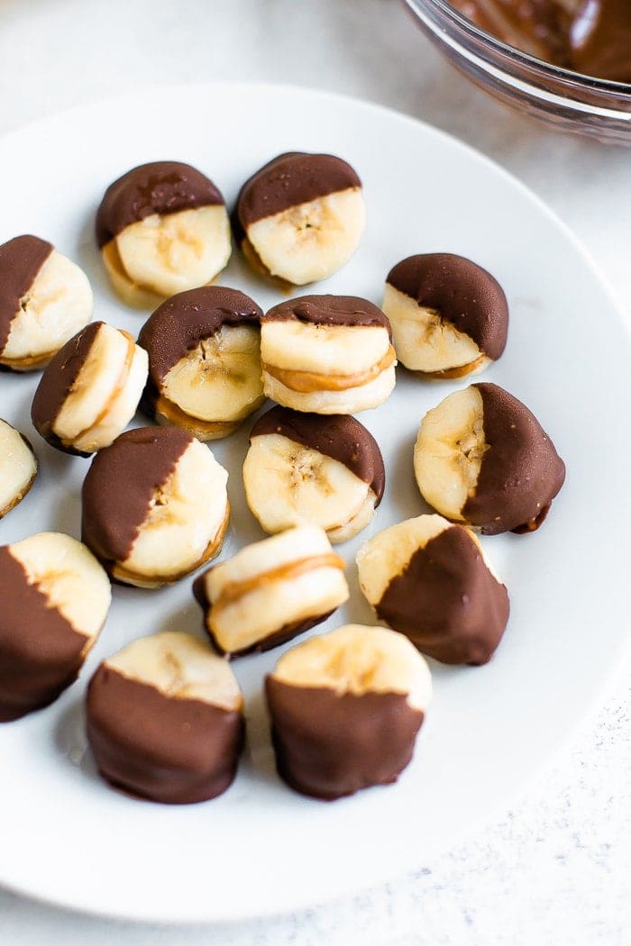 Chocolate peanut butter banana bites on a plate. Slices of banana sandwiched with peanut butter and dipped half in chocolate and frozen.