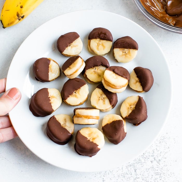 Chocolate peanut butter banana bites on a plate. Slices of banana sandwiched with peanut butter and dipped half in chocolate and frozen.