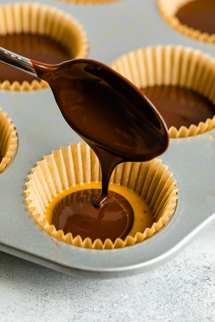 Spoon drizzling the top layer of chocolate onto a homemade peanut butter cup in a muffin tin with liners.