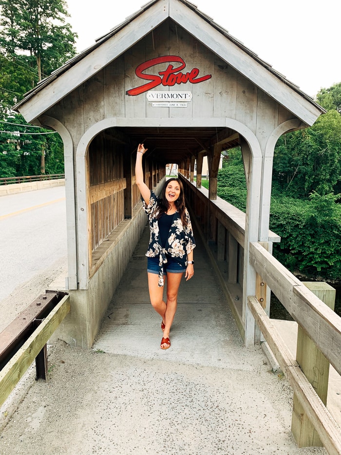 Woman standing and waving under a covered bridge that has a Stowe Vermont sign on the top.