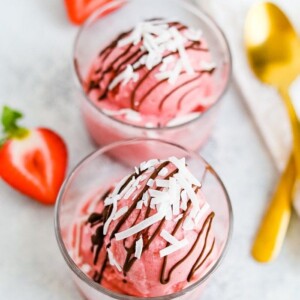 Two glasses with a scoop of strawberry coconut milk sorbet drizzled with chocolate and coconut flakes. Two spoons and strawberries are to the side.