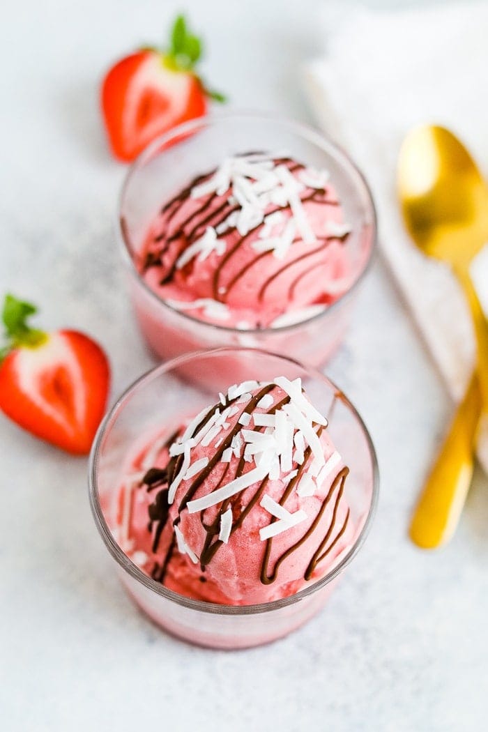 Two glasses with a scoop of strawberry coconut milk sorbet drizzled with chocolate and coconut flakes. Two spoons and strawberries are to the side.