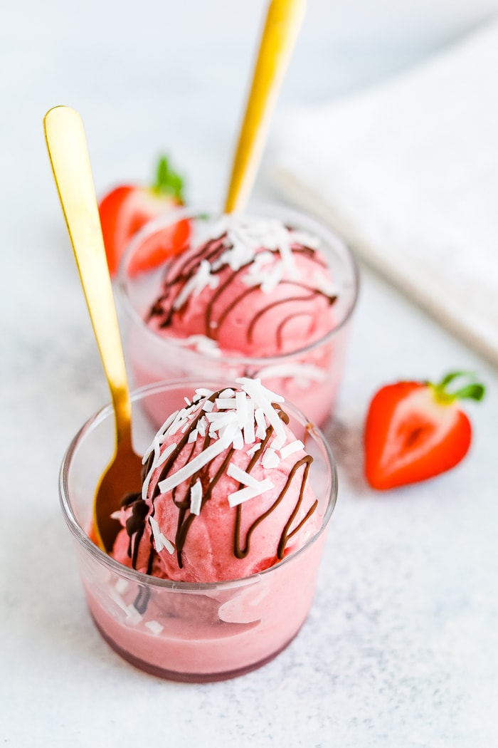 Two glasses with scoops of strawberry coconut milk sorbet with gold spoons. Sorbet is drizzled with chocolate and topped with coconut flakes.