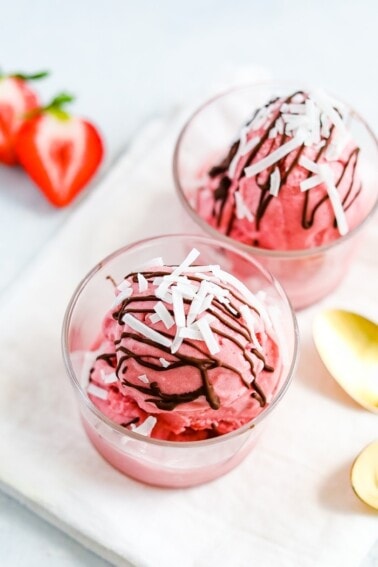 Two glasses with scoops of strawberry coconut milk sorbet with gold spoons. Sorbet is drizzled with chocolate and topped with coconut flakes.