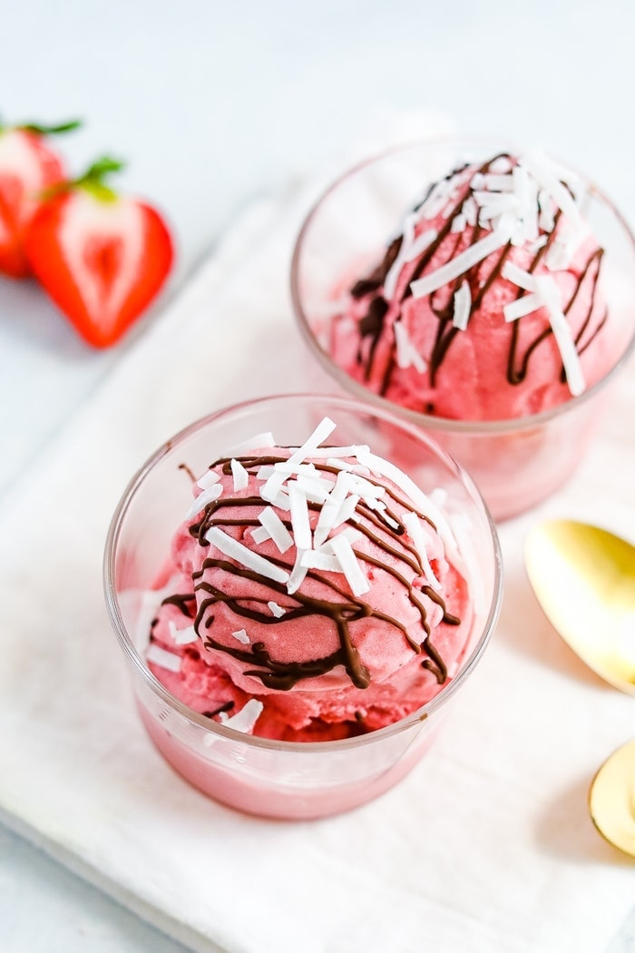 Two glasses with a scoop of strawberry coconut milk sorbet drizzled with chocolate and coconut flakes. Two spoons and strawberries are to the side.