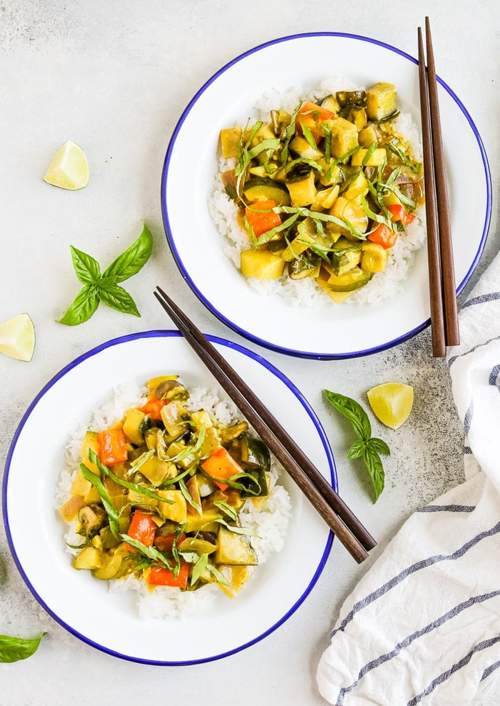 Two plates with summer vegetable coconut curry served over rice. Topped with basil and chop sticks are on the side of the plates. Fresh basil and limes are around the two plates.