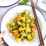 Hand holding a plate of summer vegetable coconut curry served over rice and topped with basil. Two chop sticks are on the side of the bowl.