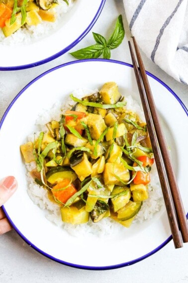 Hand holding a plate of summer vegetable coconut curry served over rice and topped with basil. Two chop sticks are on the side of the bowl.