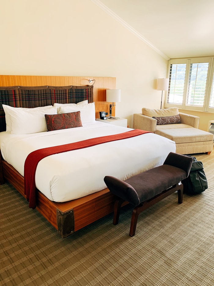 Hotel resort bedroom with a neatly made bed, bench, and chair next to a window.