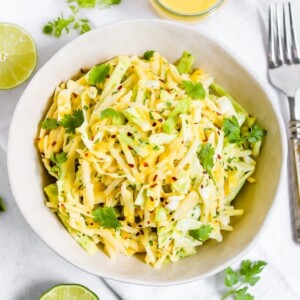 Jicama slaw in a bowl with lime halves, cilantro, dressing and a fork on the side.